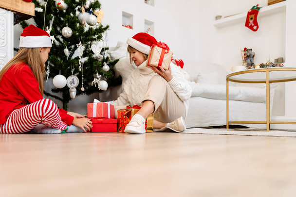 White girl and her grandmother sitting with gift boxes by Christmas tree at home - Fotografie, Obrázek