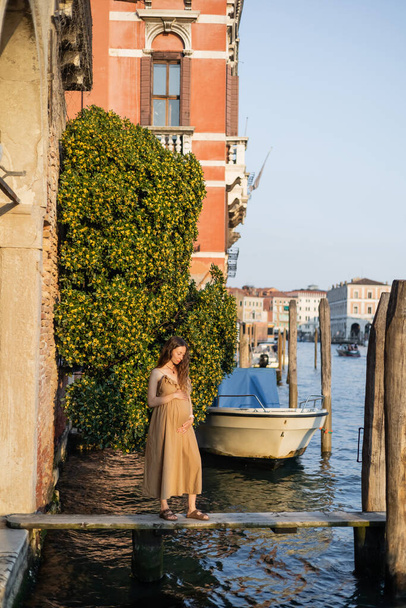 Вагітна жінка в одязі торкається живота на дерев "яному пірсі у Венеції.  - Фото, зображення