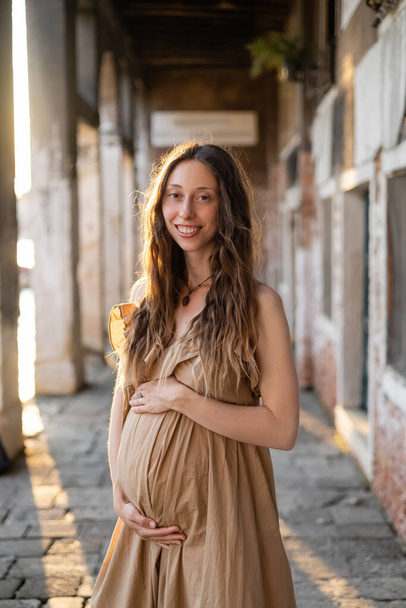 Femme enceinte positive en robe regardant la caméra près de vieux bâtiment flou en Italie  - Photo, image