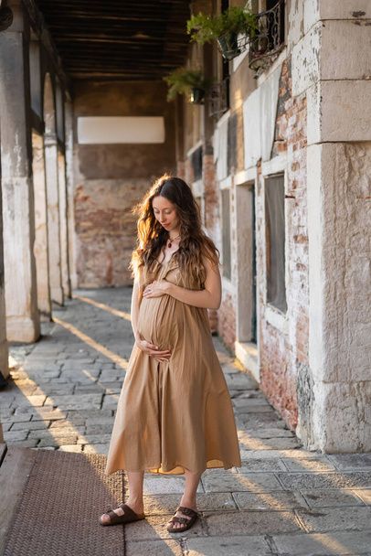 Pregnant woman in summer dress touching belly on urban street in Venice  - Photo, image