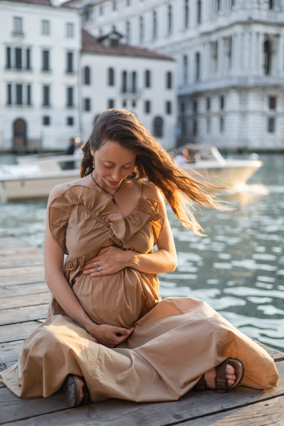 Smiling pregnant woman sitting on wooden pier in blurred Venice  - Фото, зображення