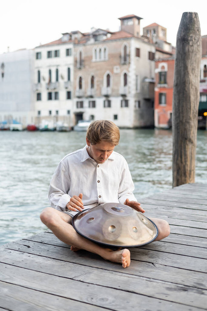 Босоногий музыкант играет на висячем барабане на пирсе возле реки в Венеции  - Фото, изображение