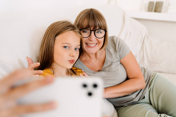 White girl and her grandmother taking selfie on cellphone while sitting on sofa at home - Foto, immagini