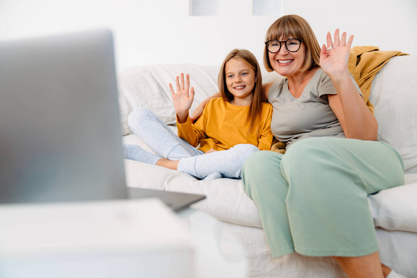 White girl and her grandmother gesturing while using laptop on sofa at home - Foto, afbeelding