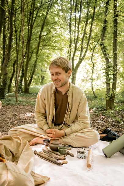 Smiling man sitting near aroma stick and teapot during tea ceremony in forest  - Valokuva, kuva
