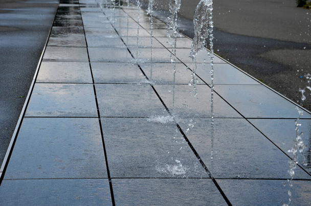 quadratische Granitpflastersteine gestapelt in einem Streifen auf dem Bürgersteig, Silos, Wasserspritzer mit wechselnden Sprühhöhen. . Wasserspiele entstehen direkt aus dem gepflasterten Platz in der Parkreihe. Nasse Fliesen auf der Promenade - Foto, Bild