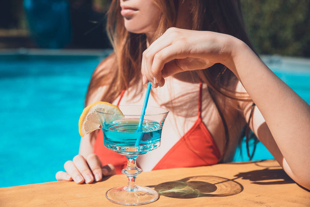 Красивая девушка-подросток в бикини пьет синий коктейль с соломинкой в баре у бассейна - Фото, изображение
