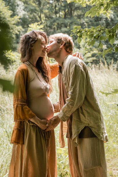 Vista lateral do homem beijando sorrindo esposa grávida na floresta  - Foto, Imagem