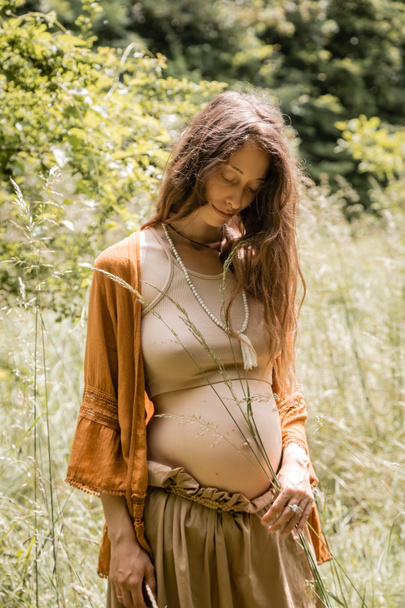 Brunette pregnant woman holding grass near belly in forest  - Foto, Imagen