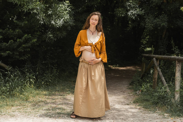 Pregnant woman looking at camera while standing on path in summer forest - Foto, Imagem