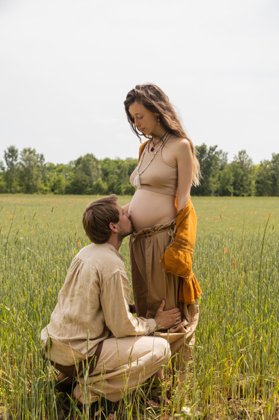 Seitenansicht des Mannes küsst Bauch der schwangeren Frau in grasbewachsenem Feld  - Foto, Bild