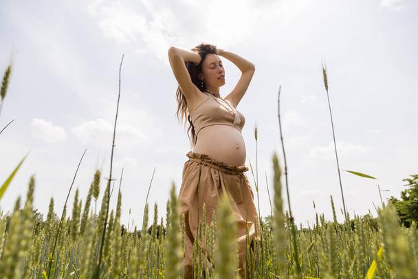 Low angle view of pregnant woman touching hair in field with spikelets - Foto, Imagen