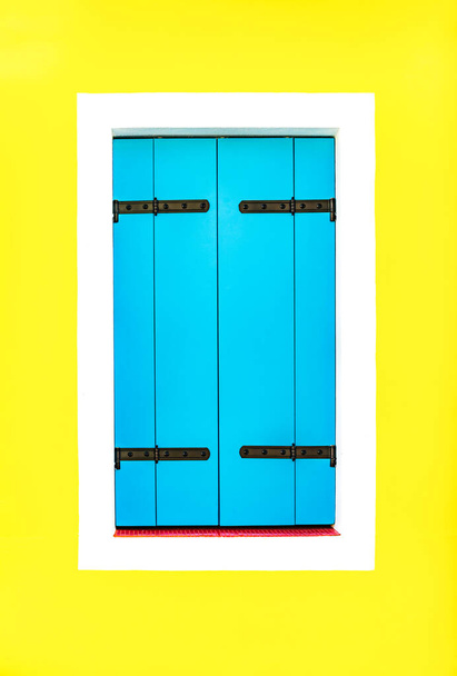 Jedno okno s bílým okrajem je zakryté dřevěnými modrými okenicemi proti jasně žluté malované stěně - Fotografie, Obrázek