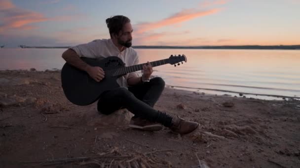 Uomo caucasico spagnolo che suona la chitarra sulla spiaggia vicino al lago al tramonto - Video 4K orizzontale - Filmati, video
