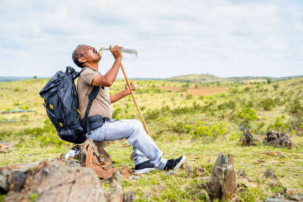 Spragniony człowiek pijący wodę siedząc na szczycie wzgórza podczas trekkingu - koncepcja robienia przerwy, zdrowego aktywnego stylu życia i hobby - Zdjęcie, obraz