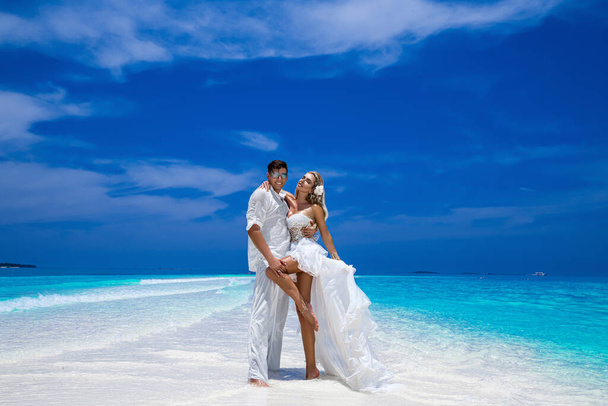 夏の愛。結婚式の服を着た美しい幸せな若いカップルがモルディブのビーチに立っています。モルディブのパラダイス島のビーチでの婚約と結婚式。高級旅行. - 写真・画像