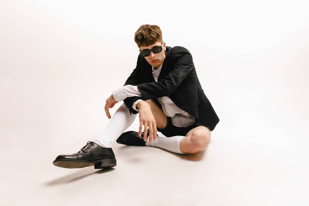 Look. Wide angle view of young fashionable man, student sitting on floor isolated on white studio background. Concept of art, beauty, fashion, youth culture, sales and ads. Looks composed, confidently - 写真・画像