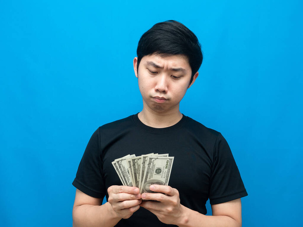 Азиатский мужчина смотрит на деньги в руке и испытывает сомнения синий фон - Фото, изображение