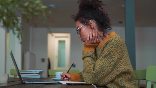 Happy woman wearing yellow sweater and eyeglasses writing something in notepad from laptop and drinking coffee in cafe - Footage, Video