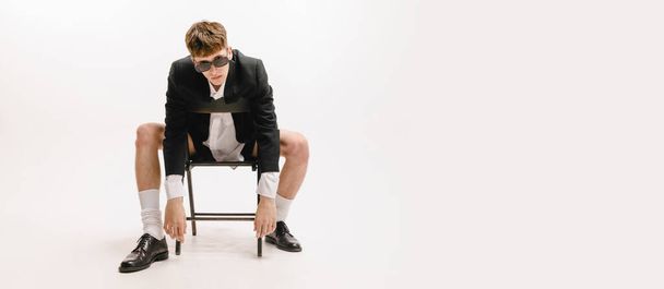 Self-confident young man in sunglasses sitting on chair and looking at camera isolated over white background. Modern business style clothes. Concept of art, beauty, fashion, youth culture, ads - Photo, Image
