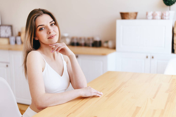 Die junge Frau sitzt am Holztisch vor der weißen Küche. Mädchen blickt vorsichtig in die Kamera. Wohnungen mit guter Reparatur mieten. - Foto, Bild