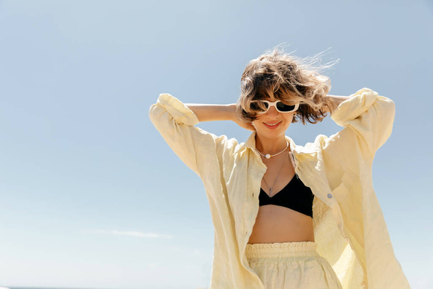 Stylish adorable pretty woman with wavy hair wearing linen yellow suit in sunglasses dancing and having fun against blue sky in summer sunny day - Φωτογραφία, εικόνα
