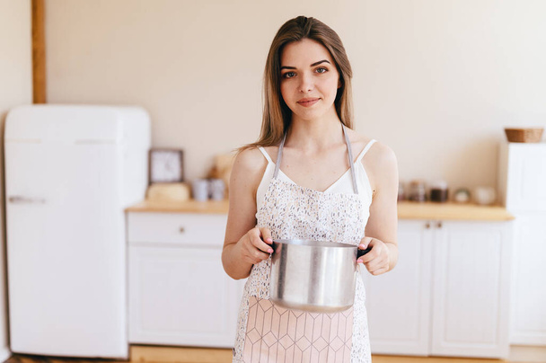 Молода жінка тримає горщик в руках і носить фартух, готуючи смачне блюдо. Світла затишна кухня у фоновому режимі
. - Фото, зображення
