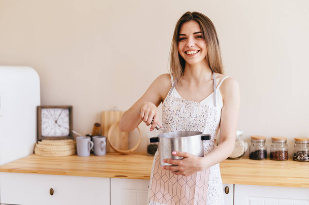 Happy Young Woman Holds Saucepan in Her Hands and Stirs with Whisk. Girl in Apron prepares delicious dish. Light Cozy Kitchen in Background - Foto, immagini