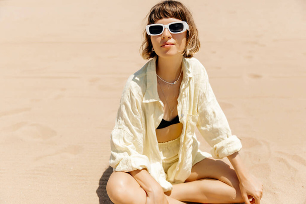 Stylish charming girl with shirt hairstyle wearing yellow summer linen suit in sunglasses resting on dandy beach in summer sunny day.  - Valokuva, kuva