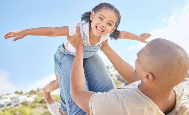 Father and daughter bonding together outdoors. Portrait of adorable little girl from below having fun pretending to fly like a superhero with arms outstretched while being held by her loving dad - 写真・画像