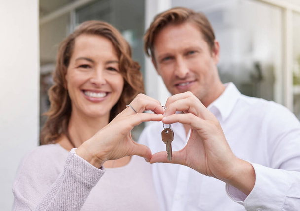 Closeup hands of happy married couple holding up new house keys with a heart shape hand gesture. Man and woman holding keys to new house or apartment - Foto, imagen