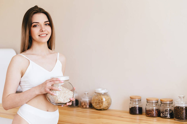 Young Woman Stands Against Background of Kitchen and Holds Jar of Oatmeal in Her Hands. Girl in white underwear in Beige Kitchen. Cozy Home Interior. - Valokuva, kuva