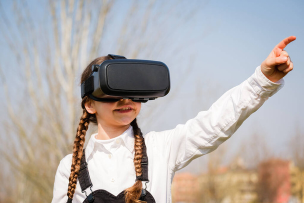 Little girl with virtual reality glasses. Future technology concept - Beautiful girl with virtual reality outdoor in a park - Photo, image