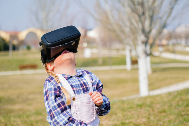 Klein meisje met een virtual reality bril. Toekomstige technologie concept - Mooi meisje met virtual reality outdoor in een park - Foto, afbeelding