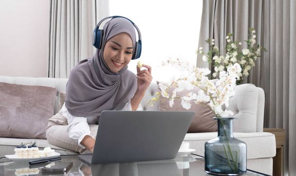 Happy Arab housewife in hijab relaxing with sweets and coffee on sofa at home enjoy indoor relaxation Relax on the comfy sofas, free space - Photo, image
