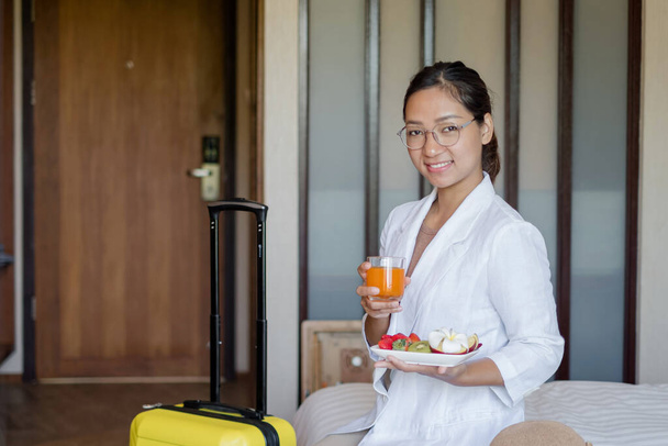 Una mujer de negocios asiática se sienta en una cama en una habitación de hotel, agotada de un viaje de negocios, lista para recibir un bocadillo de frutas del hotel, arreglado para huéspedes VIP. - Foto, imagen