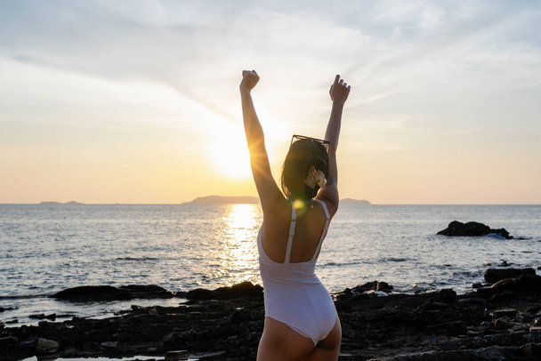 Nő turista felemeli a kezét naplementével a tengerparton nyaralni. Fürdőruhát viselő nő pihentető és felemelt karokkal, homokszemekkel a bőrén. Ázsiai hölgy élvezi a természetet nyaralni. Szabadság fogalma - Fotó, kép