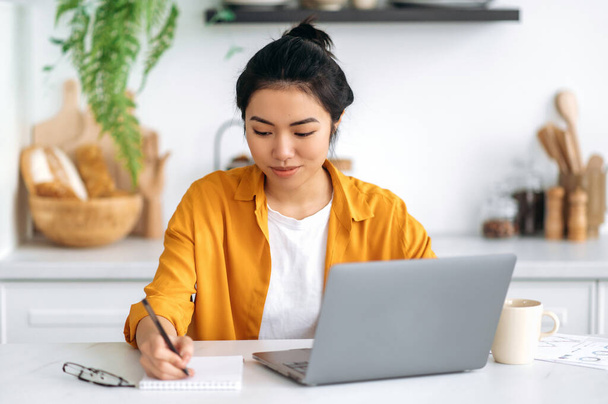 Positive pretty Chinese girl, student, freelancer, creative designer, sitting at home in the kitchen at a table with a laptop, working or studying online, working on a project, doing homework, smiling - Zdjęcie, obraz