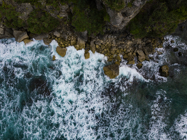 Вид сверху Голубоватые океанские волны, разбивающиеся о скалы на пляже, производят белую рябь и пену на тропическом пляже - Фото, изображение