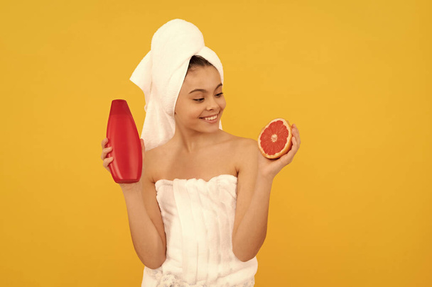 fröhliche Teenager-Kind im Handtuch mit Grapefruit-Shampoo-Flasche auf gelbem Hintergrund. - Foto, Bild