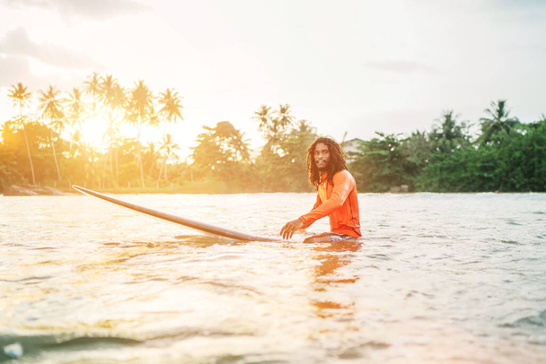 Black long-haired teen man floating on long surfboard, waiting for a wave ready for surfing with palm grove litted sunset rays. Extreme water sports and traveling to exotic countries concept. - Fotoğraf, Görsel