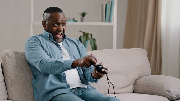 Excited worried emotional adult african american male gamer sitting on sofa in living room playing video game enjoys hobby holding controller joystick plays computer online games play console gaming - Foto, Imagen