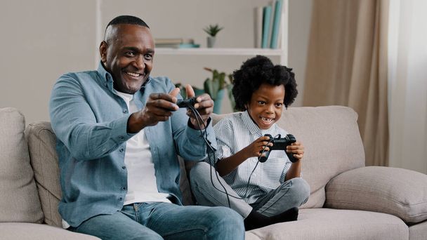 Happy african american family dad and daughter have fun together father playing video game on console with kid girl use controller sit on couch enjoying leisure activity at home winning competition - Foto, afbeelding