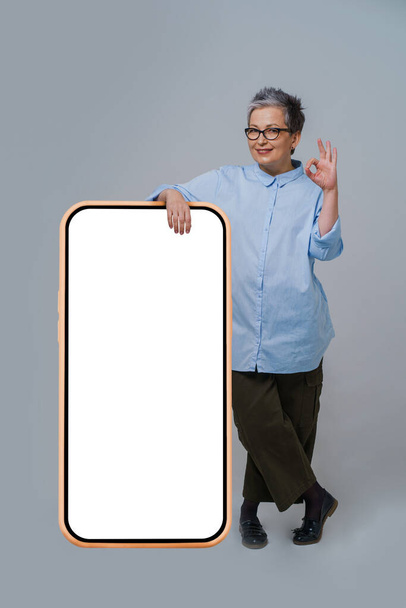 Gesturing OK grey hair mature business woman in glasses leaned on giant, huge smartphone with white screen wearing oversized blue shirt isolated on white background. Free space mock up. - Фото, зображення