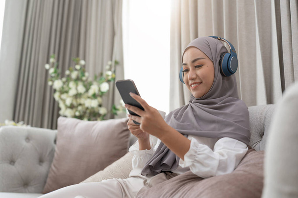Portré gyönyörű ázsiai muszlim nő alvóruhában néz online történet a mobiltelefon, lites az ágyon, és csatlakozik a vezeték nélküli internet. Fiatal aranyos lány hidzsáb hallgatni zenét okostelefon - Fotó, kép