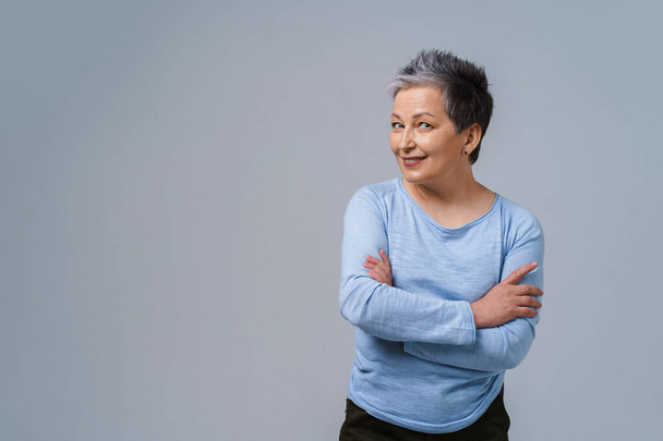 Challenging look positive emotions mature grey hair woman posing with hands folded looking at camera wearing blue blouse, isolated on white background. Healthcare, aged beauty concept. Copy space.  - Photo, image