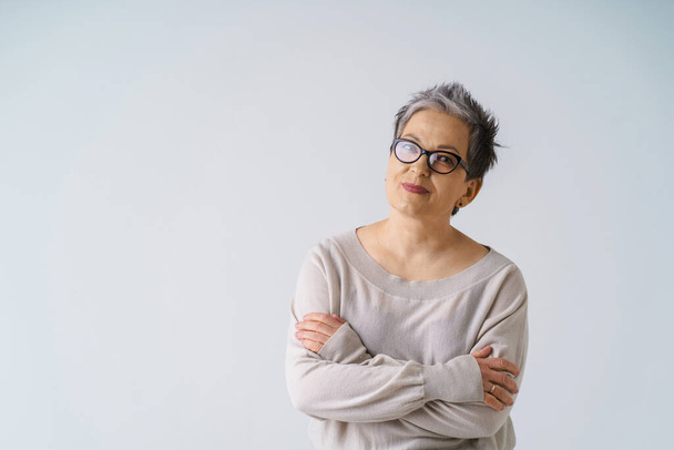Serious slightly smiling mature grey hair woman in glasses posing with hands folded looking at camera wearing white blouse, copy space isolated on white background. Healthcare, aged beauty concept.  - Foto, immagini