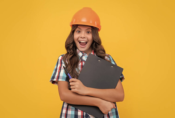 wow. überraschte Mädchen mit schützendem Hut. Erstauntes Kind im Helmhalterprojekt. Bauunterlagen. Vertragsunterzeichnung. Kindererziehung. zukünftige Ingenieure halten Klemmbrett. Sicherheitsfachkompetenz. - Foto, Bild