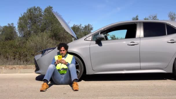 Znechucený muž, který sedí na asfaltu a píše si na mobil, stojí poblíž rozbitého auta uprostřed venkovské silnice. Porucha a oprava auta. Čeká se na pomoc - Záběry, video