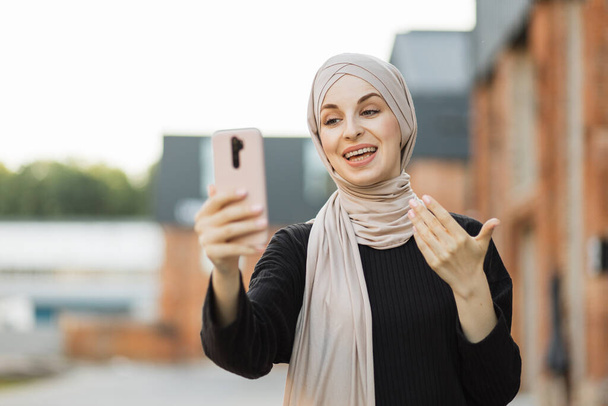 Young Muslim Arab woman in hijab takes a selfie of herself with the city background. Elegant, tall, slim and wearing black clothes and turban, smiling while takes her photo. - Φωτογραφία, εικόνα